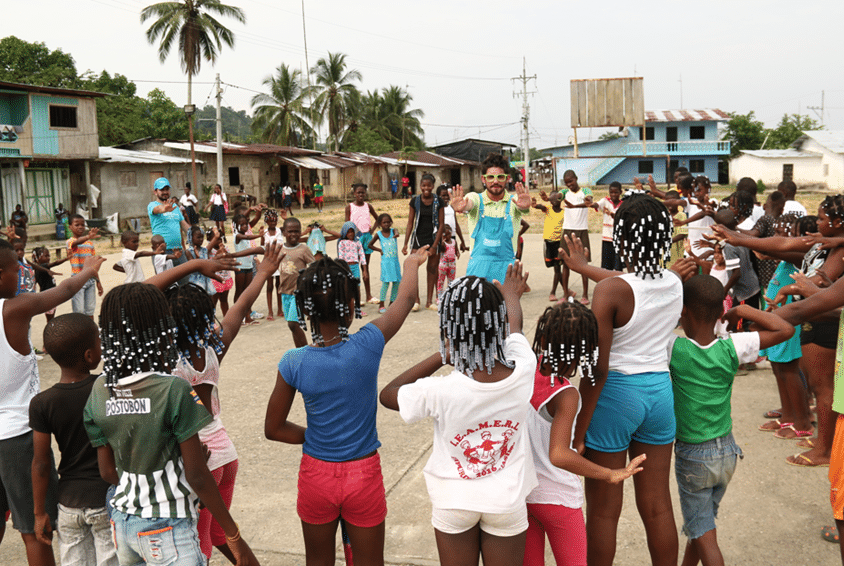SOS-barnebyer i Columbia, barna er samlet ute og leker i ring. Mange barn fra Colombia står i en ring og strekker armene i været. En medarbeider fra SOS-barnebyer med blå vest med logo, leder leken. Foto SOS-barnebyer Colombia