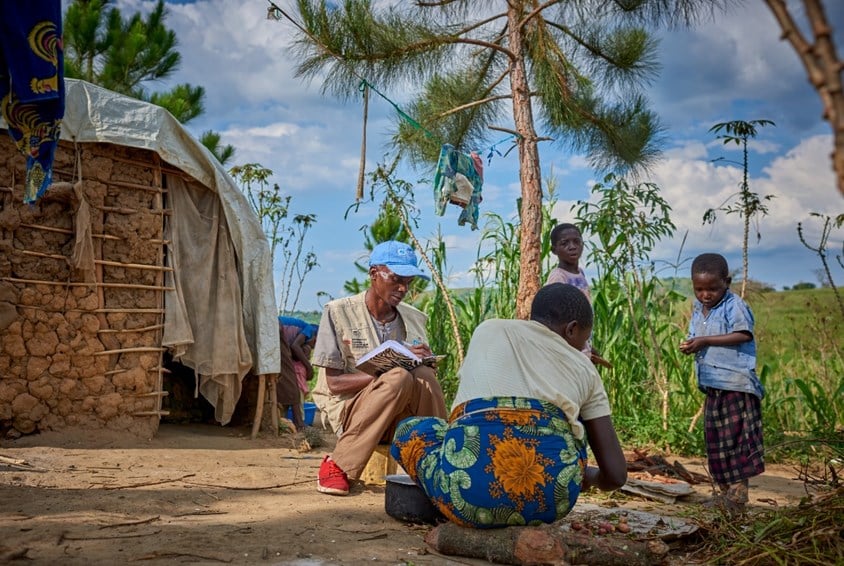 SOS-barnebyer driver flere forebyggende familieprogram i Uganda. En mor sitter på bakken og skreller grønnsaker, hun har blomstrete skjørt og lys t-skjorte, to av barna hennes står og ser på. SOS-barnebyers medarbeider sitter ved siden av. Han har blå caps, beige vest med SOS-barnebyers logo, bun bukse og røde joggesko. I bakgrunnen er huset de bor i, med vegger laget av leire og et klede foran døra. Foto: Joris Lugtigheid