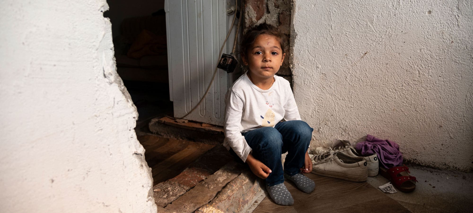 Jente med mørkt hår i hestehale, hvit genser og jeans sitter en inngang uten dør. det er slitte murvegger rundt henne. Foto: Katerina Ilievska