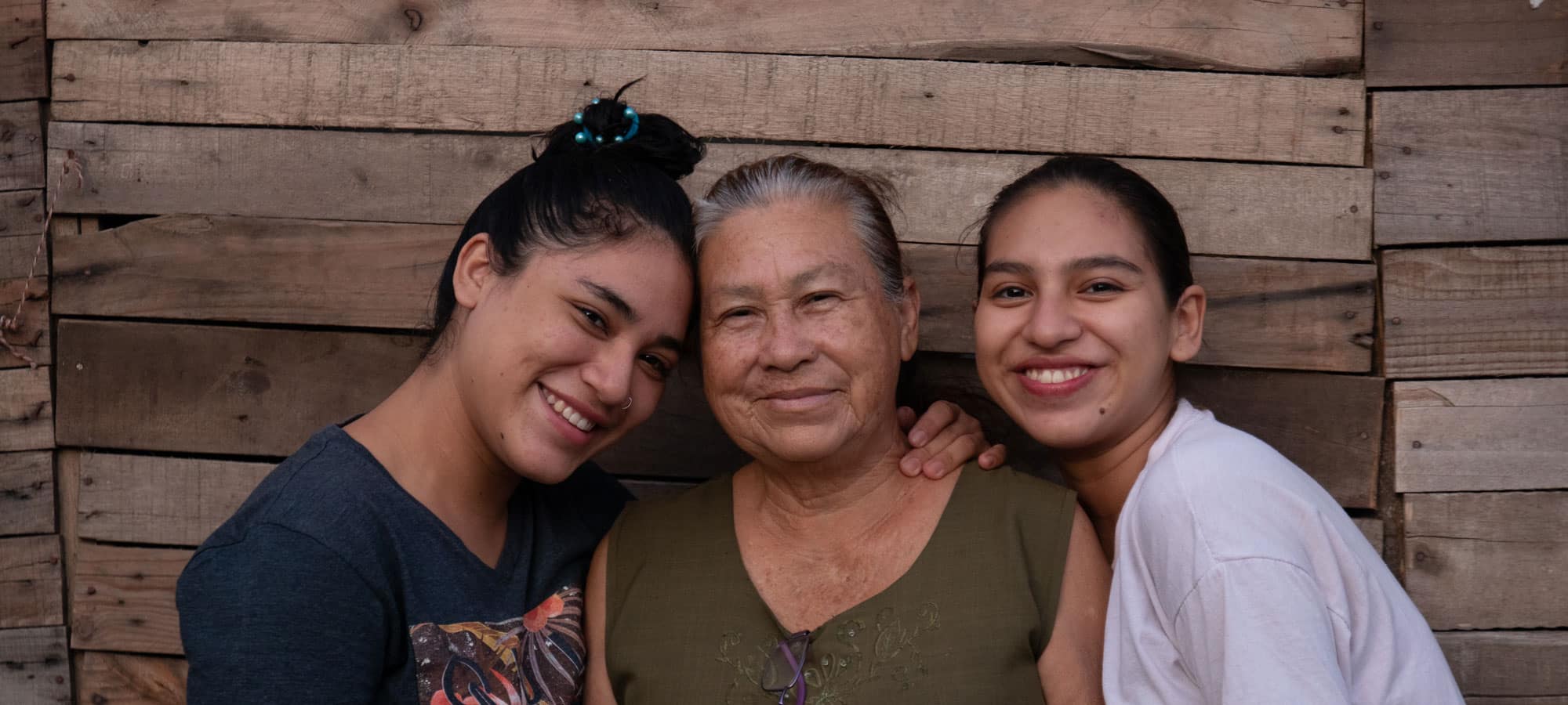 En bestemor fra Bolivia med mørkt hår i en topp, grønn t-skjorte, og lilla briller festet i halsen på t-skjorte står sammen med to barnebarn, den ene jenta har blå t-skjorte med blomster og den andre hvit. Begge smiler. Foto: Hairo Caba/Monica Garcia