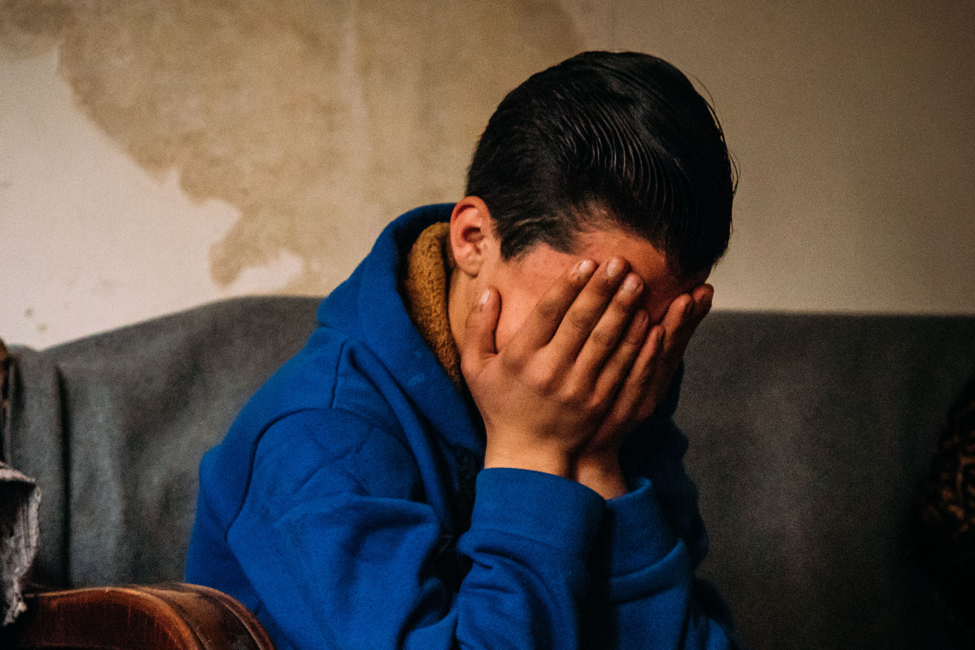 Gutt med kort, mørkt hår og blå genser sitter i en sofa og holder hendene foran ansiktet. Foto: Alea Horst 