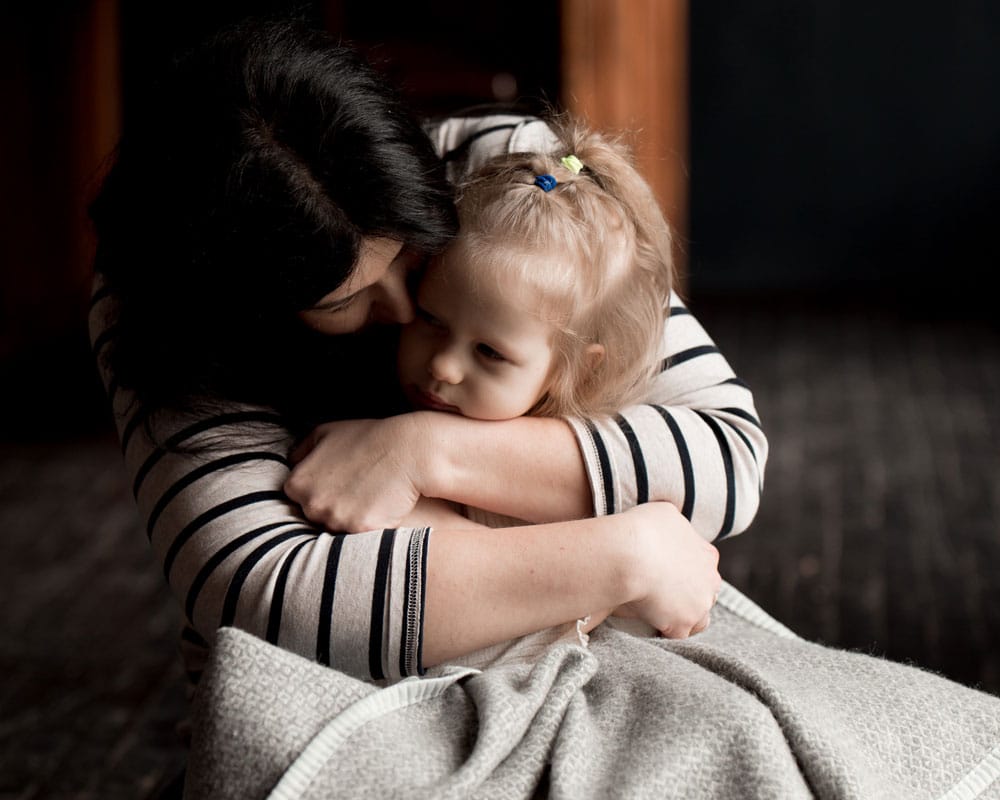 Mor med langt mørkt hår og stripete genser har datteren på fanget og holder rundt henne. hun har lyst hår med ei sløyfe og et teppe rundt seg. Foto: Katerina Ptakha