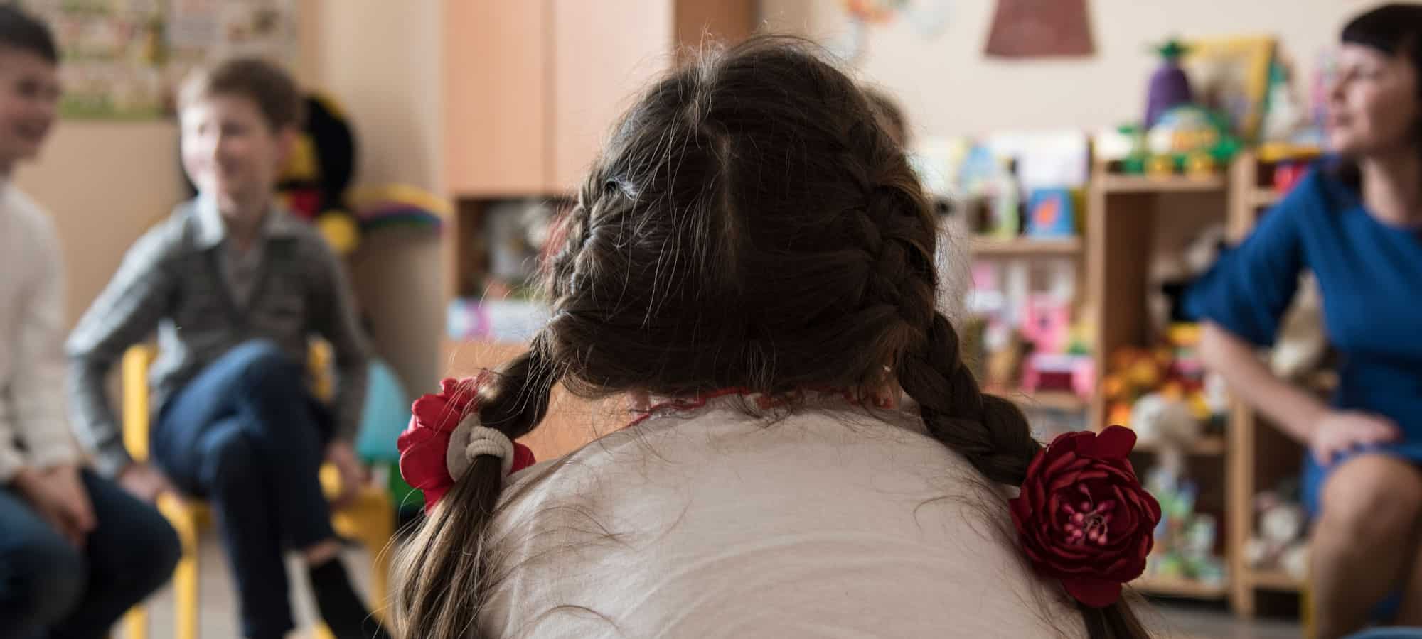 Flere barn sitter i et rom med leker og snakker med psykologen. Ei jente, med fletter og hvit genser, sitter på en stol i forgrunnen med ryggen til. Foto: Katerina Ilievska