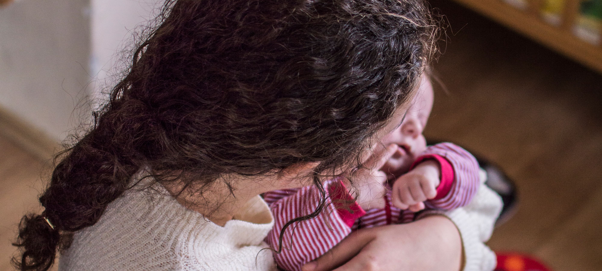 Bakhodet på en ung mor med bølgete mørkt hår i hestehale som holder en liten baby med stripete rød og hvit dress, i armene. Foto: Teodor Bø