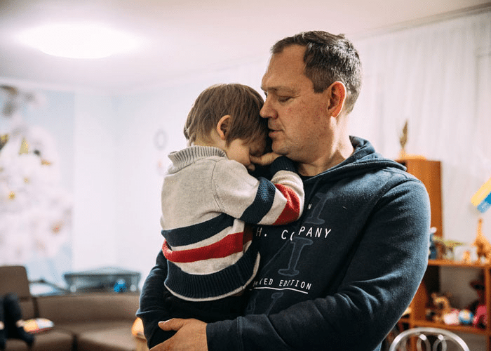 En SOS-fosterfar, med mørkeblondt kort hår og blå hettegenser, holder en liten gutt i armene, han har på seg stripete genser og mørk bukse. Foto: Alea Horst