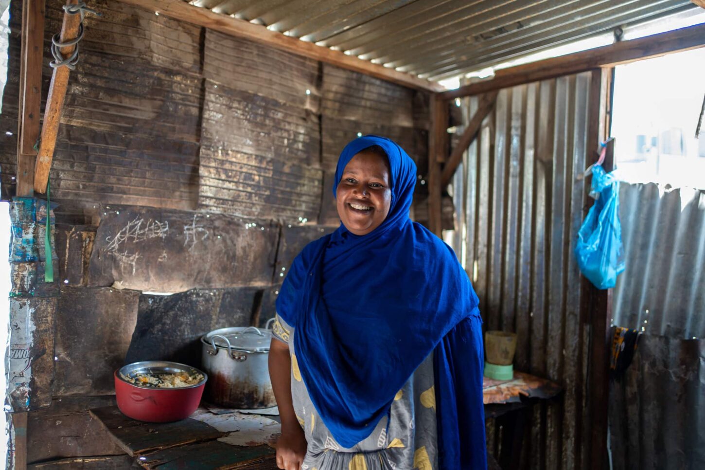 Qadan står inne i butikken sin og smiler. Hun har på seg blå hijab. På ovnsplatene står store kjeler. Foto: Lydia Mantler