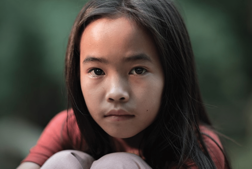 Jente fra Laos sitter med armene rundt knærne og ser alvorlig inn i kamera. Illustrasjonsfoto: Lars Just