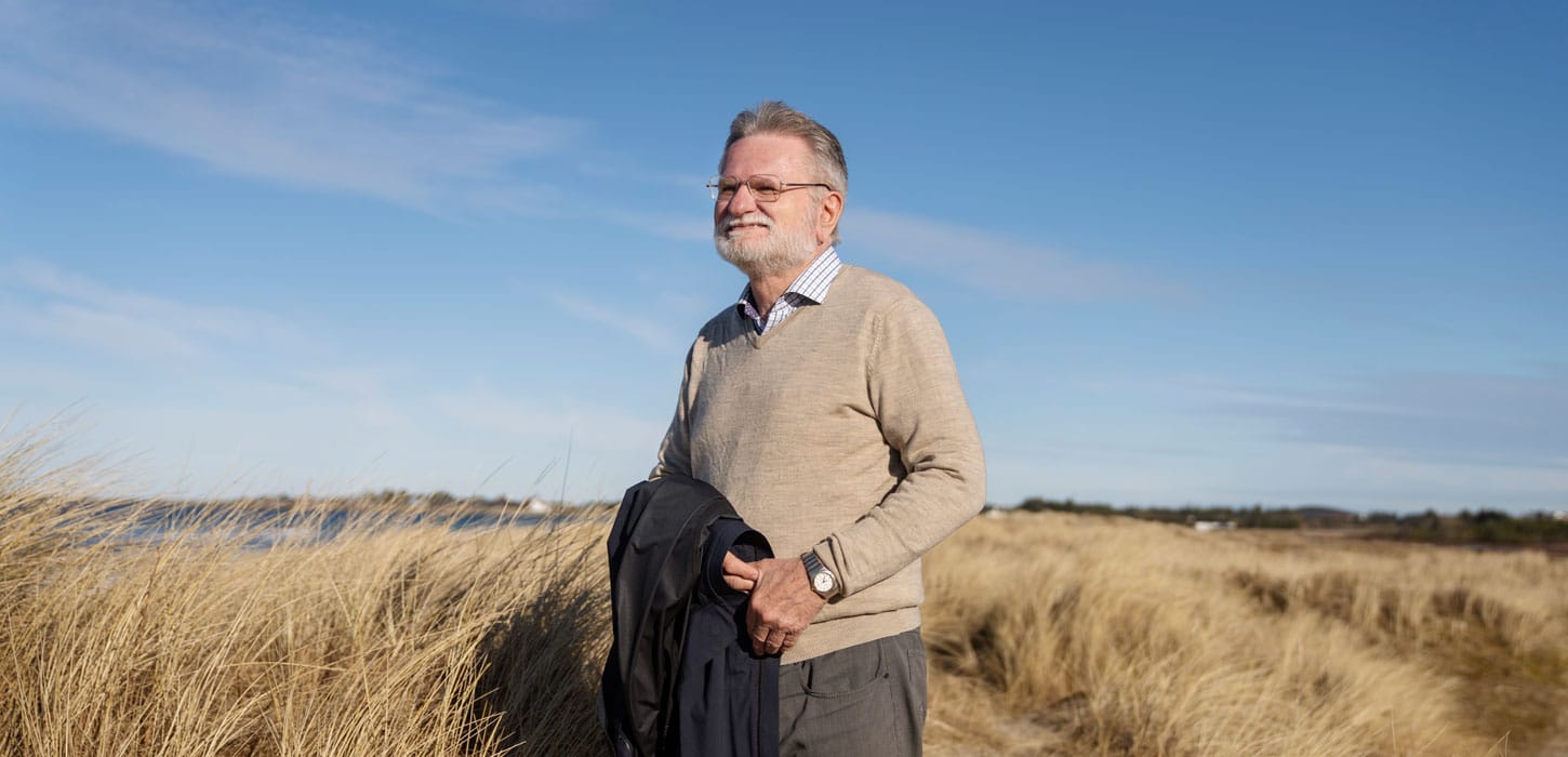 Åge Lærdal. Han har grått hår, skjegg og briller og har på seg en hvit og blå rutet skjorte, beige genser , brun bukse oghar en blå frakk hengende over armen. Han står iet landskap med høye strå, et glimt av sjø og blå himmel. Foto Marie von Krogh