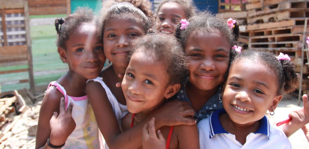En fostermor i SOS-barnebyer holder ei jente tett inntil seg og klemmer henne. Moren har mørkt langt hår i hestehale og blå t-skjorte, jenta har grønn kjole. Foto: Giti Carli Moen Seks jenter fra Colombia står tett sammen. Alle har krøllete hår med fine spenner og strikker. Foto: Foto: Giti Carli Moen