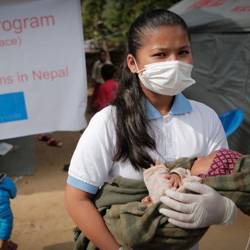 Ung kvinne, med hvit t-skjorte og langt mørkt hår i hestehale, bærer en liten baby. Bildet er fra nødhjelpsarbeid i forbindelse med jordskjelv i Nepal. Foto: Zishaan Akbar Latif