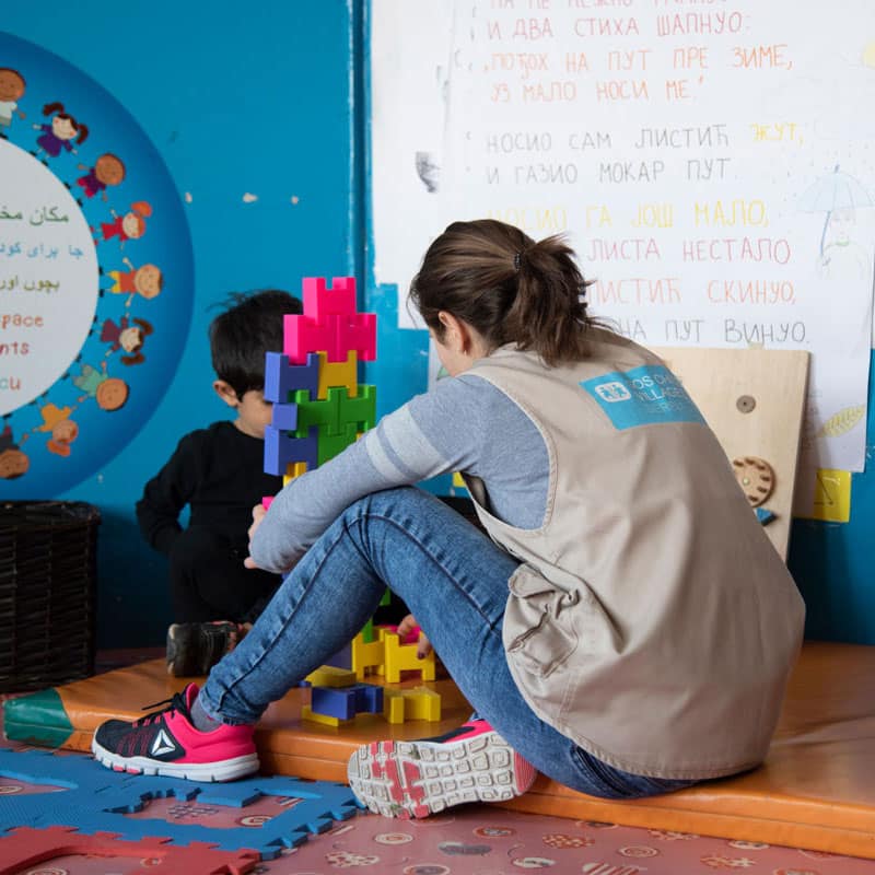 En medarbeider fra SOS-barnebyer med mørkt hår i hestehale, lyseblå genser, jeans og beite vest med SOS-barnebyers logo på ryggen sitter på gulvet med ryggen til og leker med en liten gutt med mørkt hår og mørk genser. Bildet er fra ett av SOS-barnebyers sosialsentre i Ukraina. Foto: Katerina Ilievska