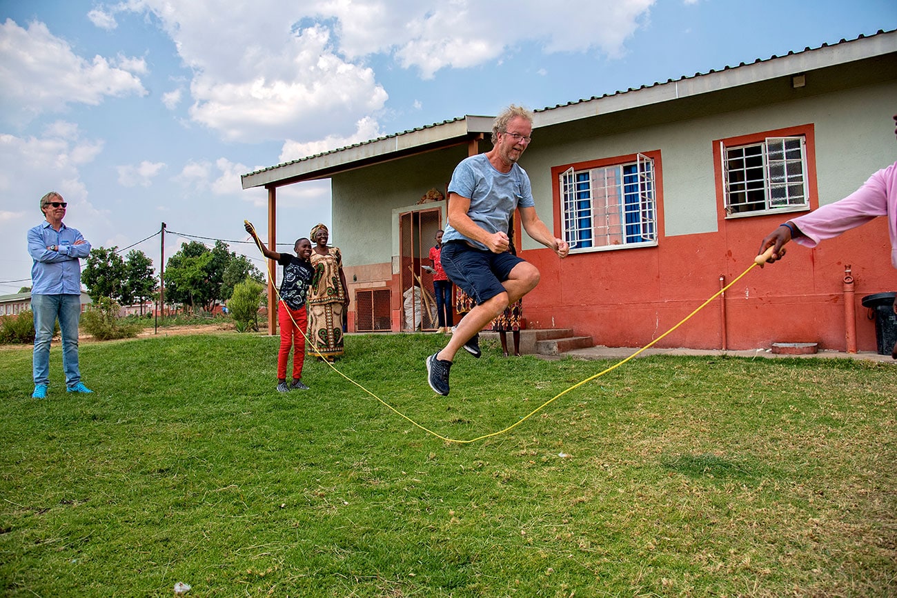 Lasse Kristiansen med lyseblå t-skjorte, mørk shorts, lyst krøllete hår, skjegg og briller hopper tau sammen med barna i Fagforbundets barneby i Huambo. I bakgrunnen er ett av husene. Foto: Ingunn Eriksen