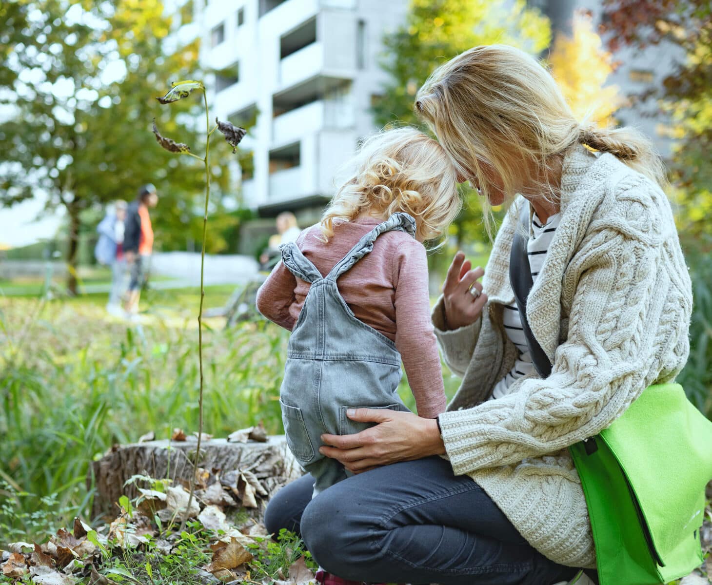 En mor med lyst, langt hår i hestehale, beige jakke og jeans, sitter på huk og holder rundt datteren sin på ca. 4 år. Hun har lyst krøllete hår, lyselilla genser og selebukser. På bakken er det løv og i bakgrunnen blokker. Illustrasjonsfoto: Gerhard Berger