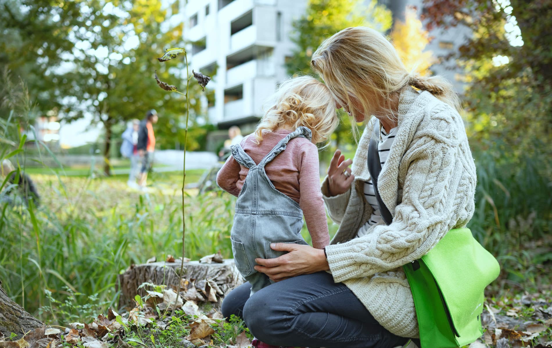 En mor med lyst, langt hår i hestehale, beige jakke og jeans, sitter på huk og holder rundt datteren sin på ca. 4 år. Hun har lyst krøllete hår, lyselilla genser og selebukser. På bakken er det løv og i bakgrunnen blokker. Illustrasjonsfoto: Gerhard Berger