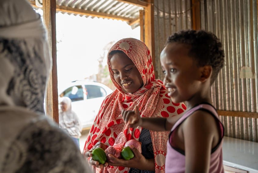 Den eldste datteren til Hodan, i rød og beige hijab, holder noen grønnsaker i hånda. ved siden av henne står den yngste broren, han har rosa singlet. Veggene i rommet er lagt av blikk. Foto: Lydia Mantler