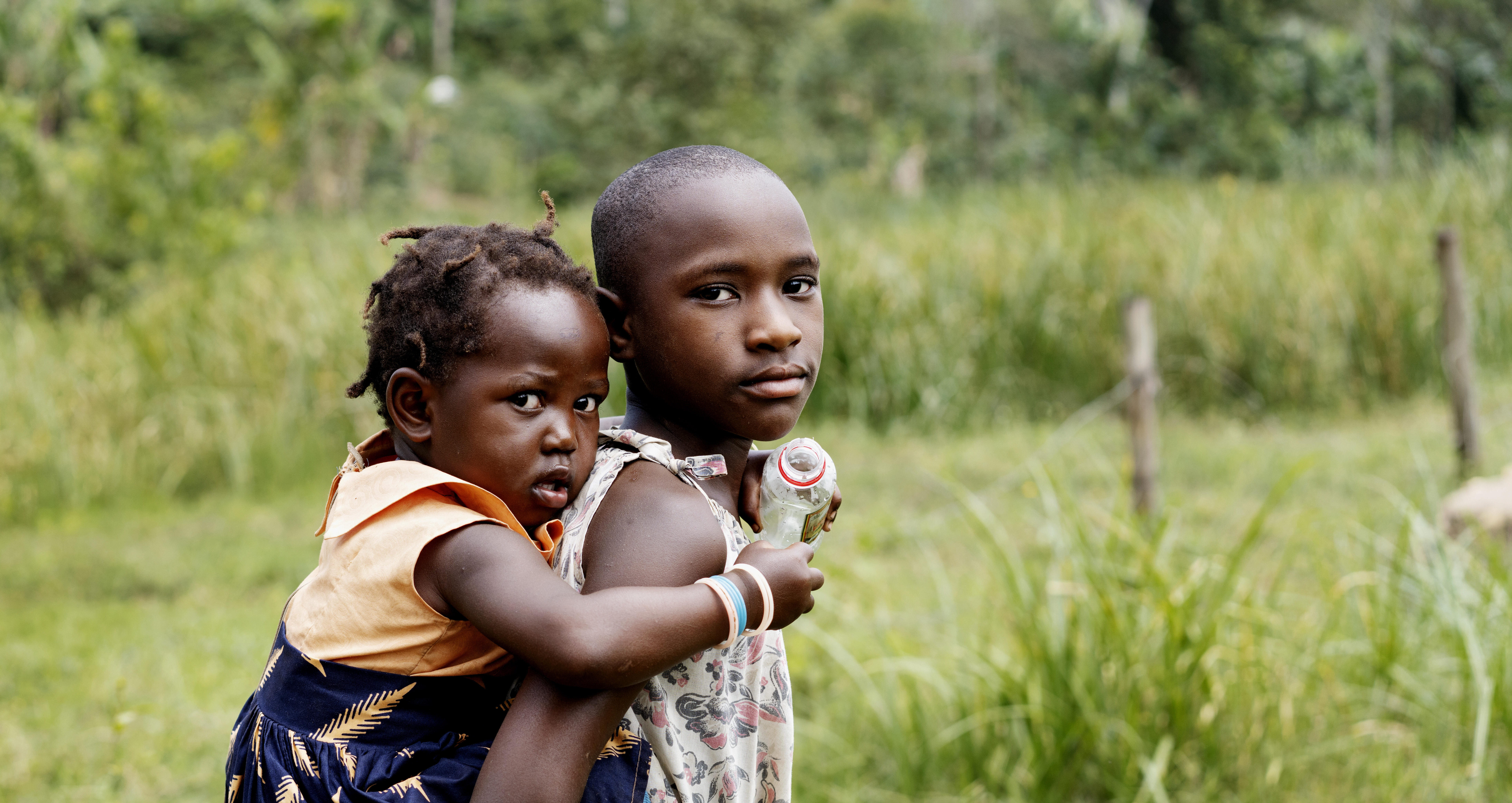 Jente bærer lillesøsteren sin på en gresslette i Uganda. Hun har på seg en beige kjole og lillesøster har på seg en blå bukse og orange trøye.