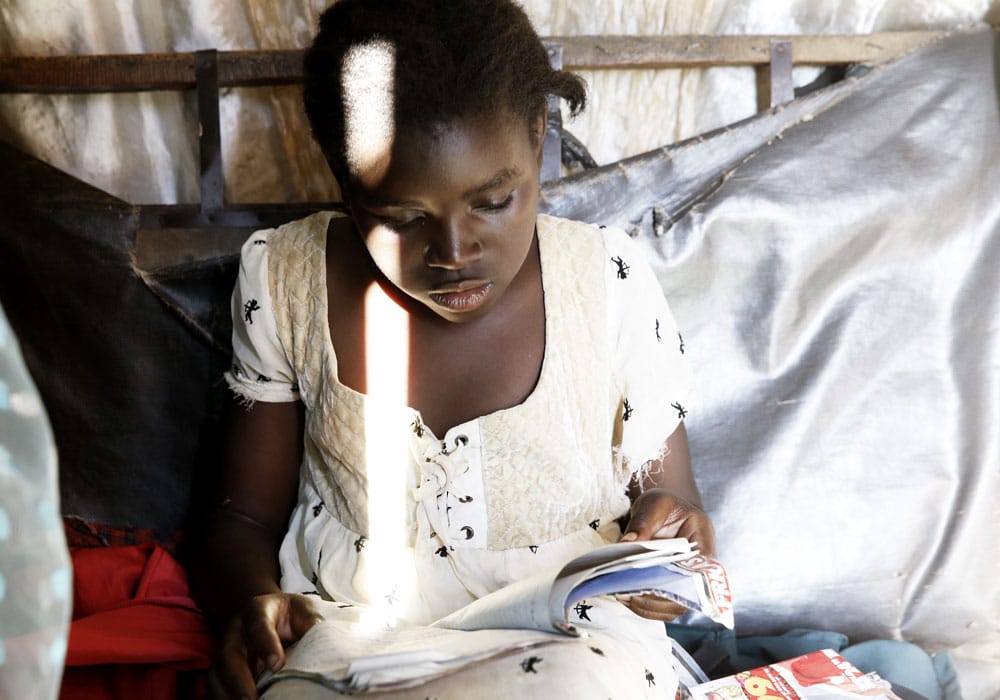 Betty med lys mønstrete kjole sitter på sengekanten og leser i ei bok. Foto: Paal Audestad