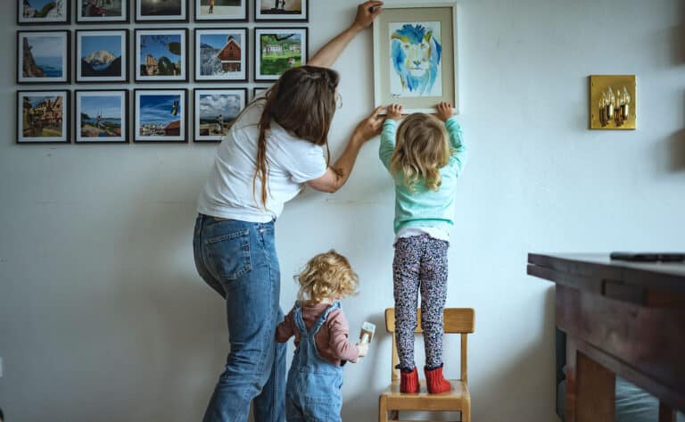 En mor og to barn, alle med ryggen til henger opp en innrammet tegning på veggen. Moren har mørkeblondt langt hår, hvit t-skjorte og jeans, den minste datteren har lyst krøllet hår, rosa genser og snekkerbukse, hun holder en hammer i hånda. Den eldste har langt bølgete hår, hun har grønn genser og mønstrete bukse, og står på en liten barnestol. Foto: Gerhard Berger