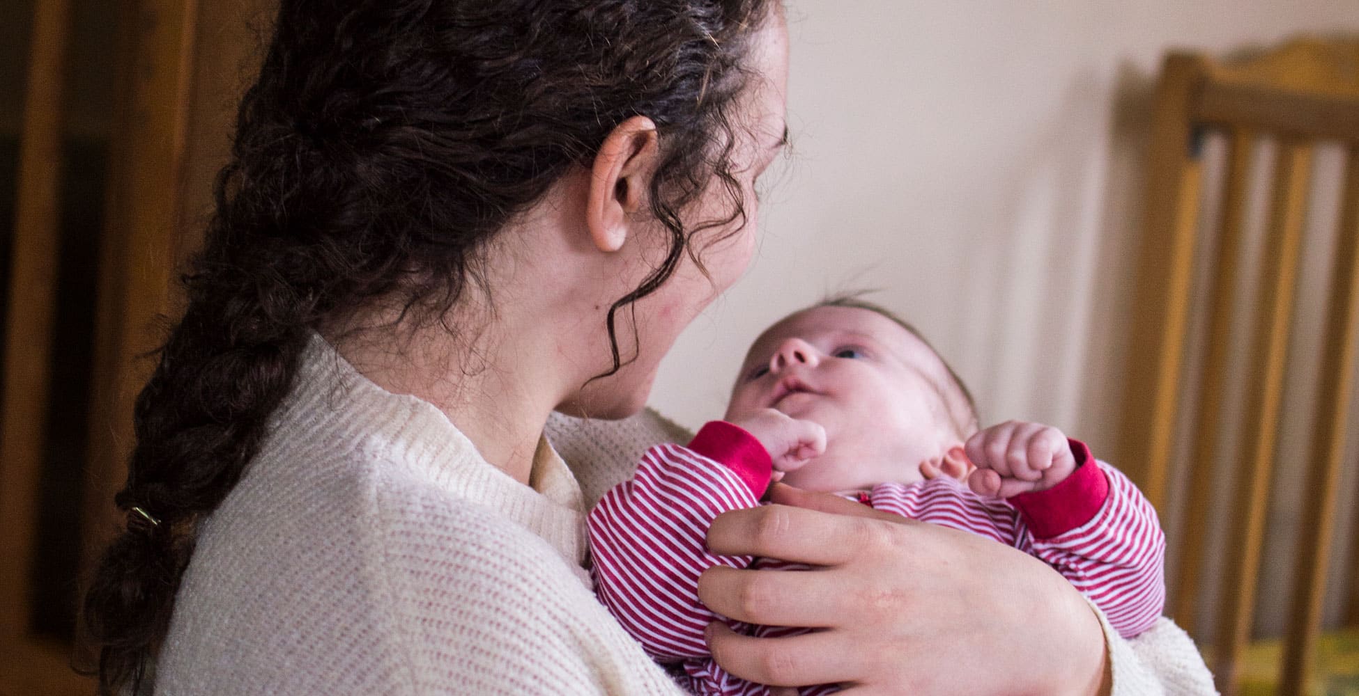 En ung mor i profil, bølgete mørkt hår i hestehale og hvit genser, holder en liten baby, med stripete rød og hvit dress, i armene. Foto: Teodor Bø