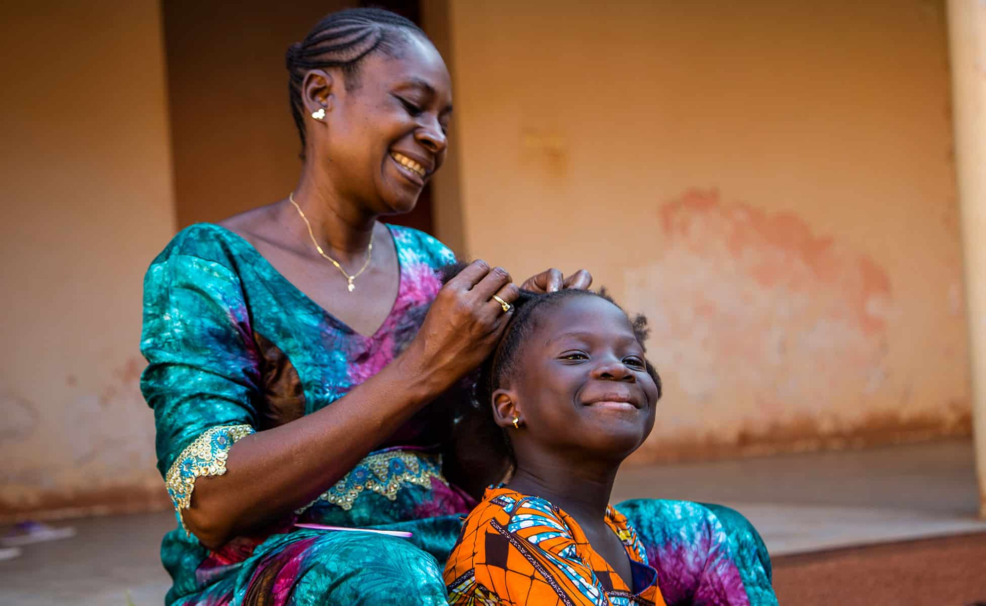 En SOS-fostermor fra et land i Vest-Afrika sitter på ei trapp og fletter håret til datteren sin. Begge har på seg fargerike klær. Foto: Sébastien Taylor