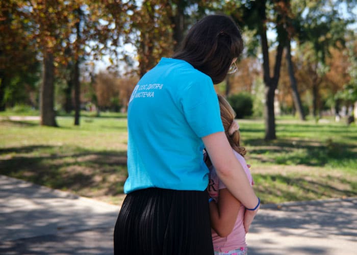 SOS-medarbeider med blå t-skjorte med SOS-barnebyers logo på ryggen holder rundt ei jente med lyst hår i hestehale, rosa strikk i håret og rosa t-skjorte. Illustrasjonsfoto Katerina Ilievska Ukraina: SOS-medarbeider med blå t-skjorte med SOS-barnebyers logo på ryggen holder rundt ei jente med lyst hår i hestehale, rosa strikk i håret og rosa t-skjorte. Illustrasjonsfoto Katerina Ilievska