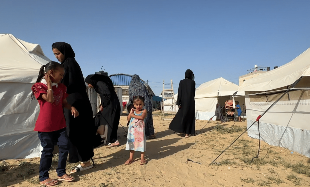 Liten jente i rosa og blå kjole som ser i kamera mellom telt i teltleir i Gaza. Juni 2024
