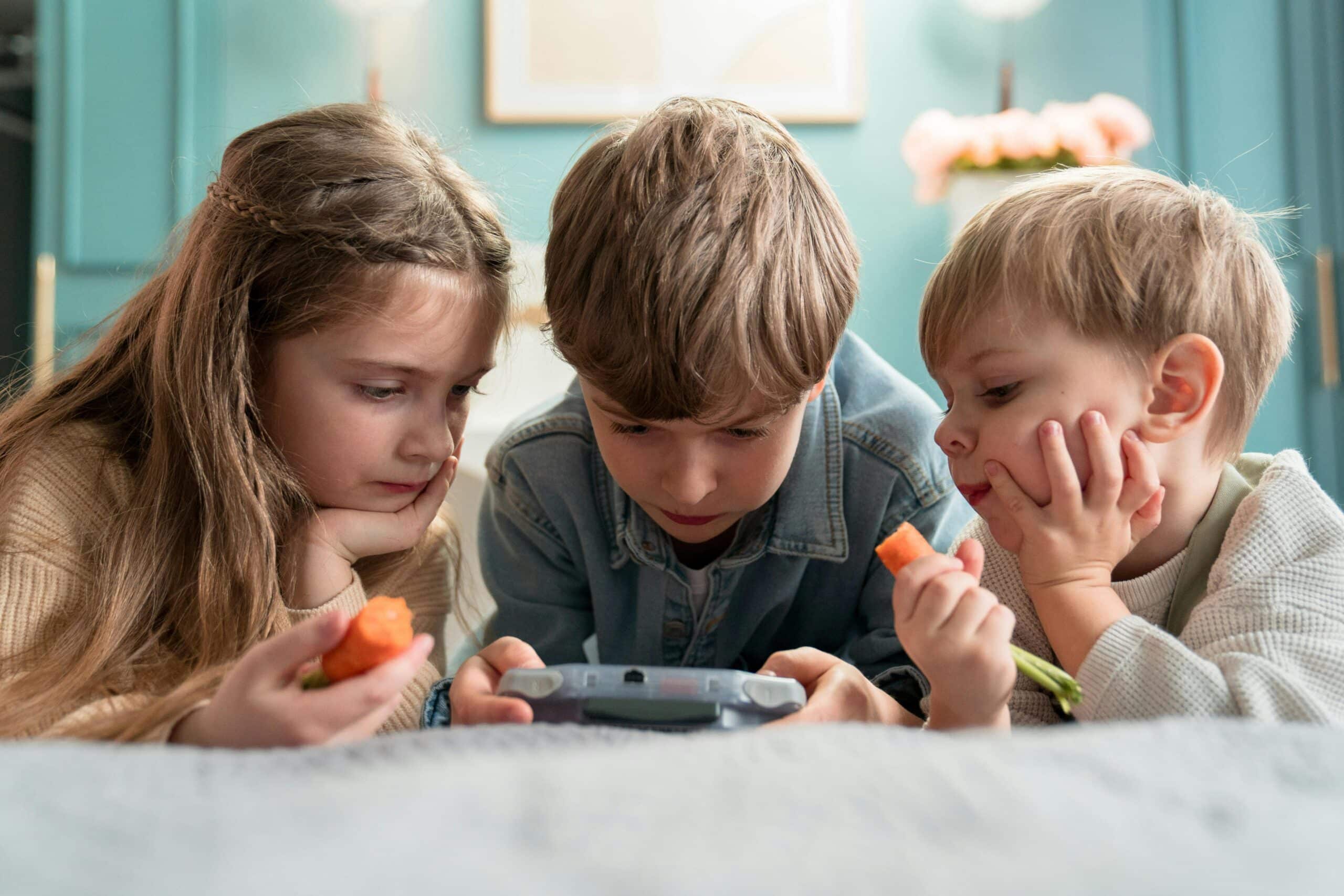 Tre søsken, to gutter med kort, mellombrunt hår og en jente med langt mellombrunt hår og flettet pannelugg, ligger på magen og spiser gulerøtter og spiller på en gameboy. Jenta og den yngste gutten ser på, mens den andre broren er den som spiller. Foto Pexels Ron Lach