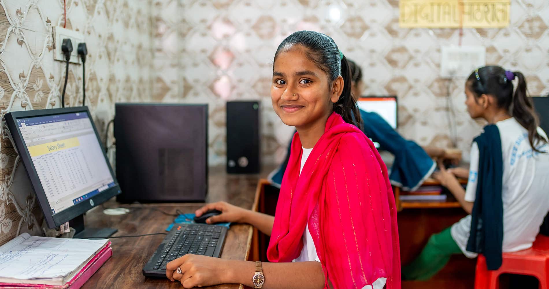Nazia, med mørkt, langt hår i hestehale, rødt langt skjerf og hvit t-skjorte med SOS-barnebyers logo, sitter foran PCen og jobber. Det er flere barn og unge i rommet som også sitter foran PCer. Foto: Lydia Mantler