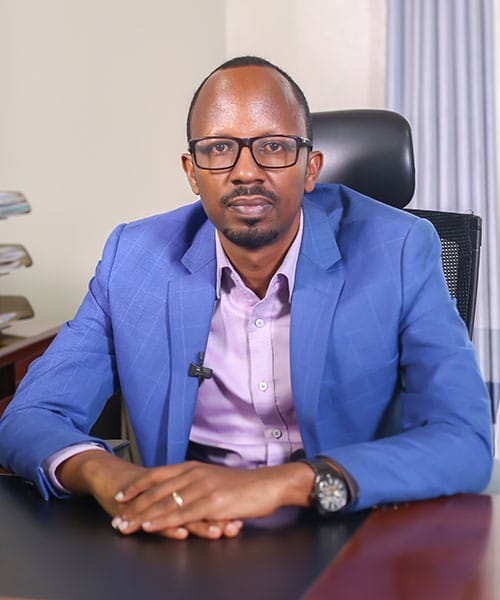 Jean Bosco Kwizera, SOS-barnebyers nasjonaldirektør i Rwanda. Han har kortklippet hår, skjegg, briller, himmelblå dress og lys lilla skjorte, og sitter på kontoret sitt med hendene foldet over PCen. Foto SOS-barnebyer