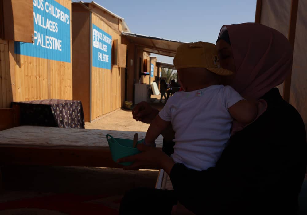 Krigen i Gaza: En mor med mørk hijab har den lille gutten sin på armen. I bakgrunnen ser vi SOS-barnebyers midlertidige leir, der husene er laget av planker. Foto Mohammad AlBaba