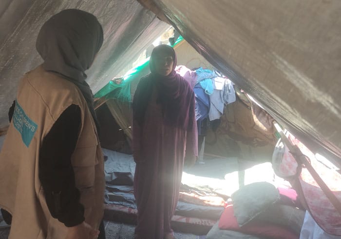 Fra en teltleir i Gaza: Kvinne med hijab og lang kjole står i silhuett i en teltåpning, foran henne med ryggen til står en av SOS-barnebyers medarbeidere. Hun har mørk hijab, kjole og beige vest med SOS-barnebyers logo på ryggen. Foto SOS-barnebyer Palestina