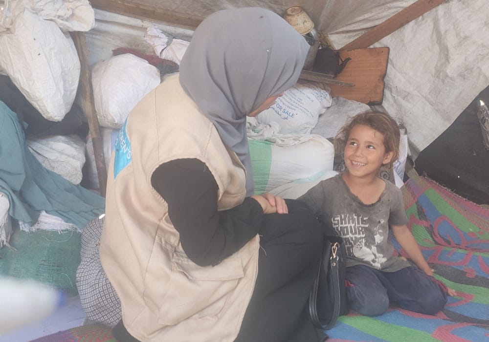 Fra en teltleir i Gaza: Ei lita jente med mørkt, bølgete hår i hestehale, grå t-skjorte og jeans sitter på gulvet i et provisorisk telt, Hun smiler til SOS-barnebyers medarbeider som har grå hijab, mørk kjole og beige vest med SOS-barnebyers logo på ryggen. Foto SOS-barnebyer Palestina