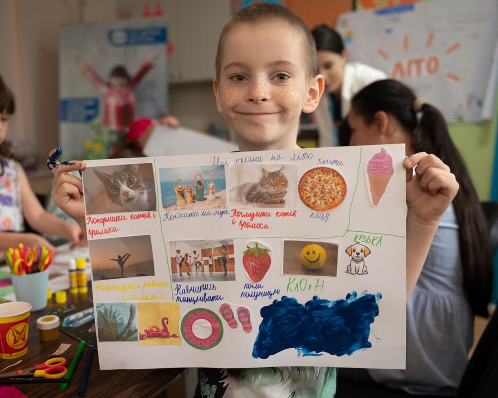 Ukraina: Artim smiler og holder opp en plakat med forskjellige tegninger og utklipp som viser alt han har lyst til å gjøre. Han har kort, blondt hår. Foto Katerina Ilievska