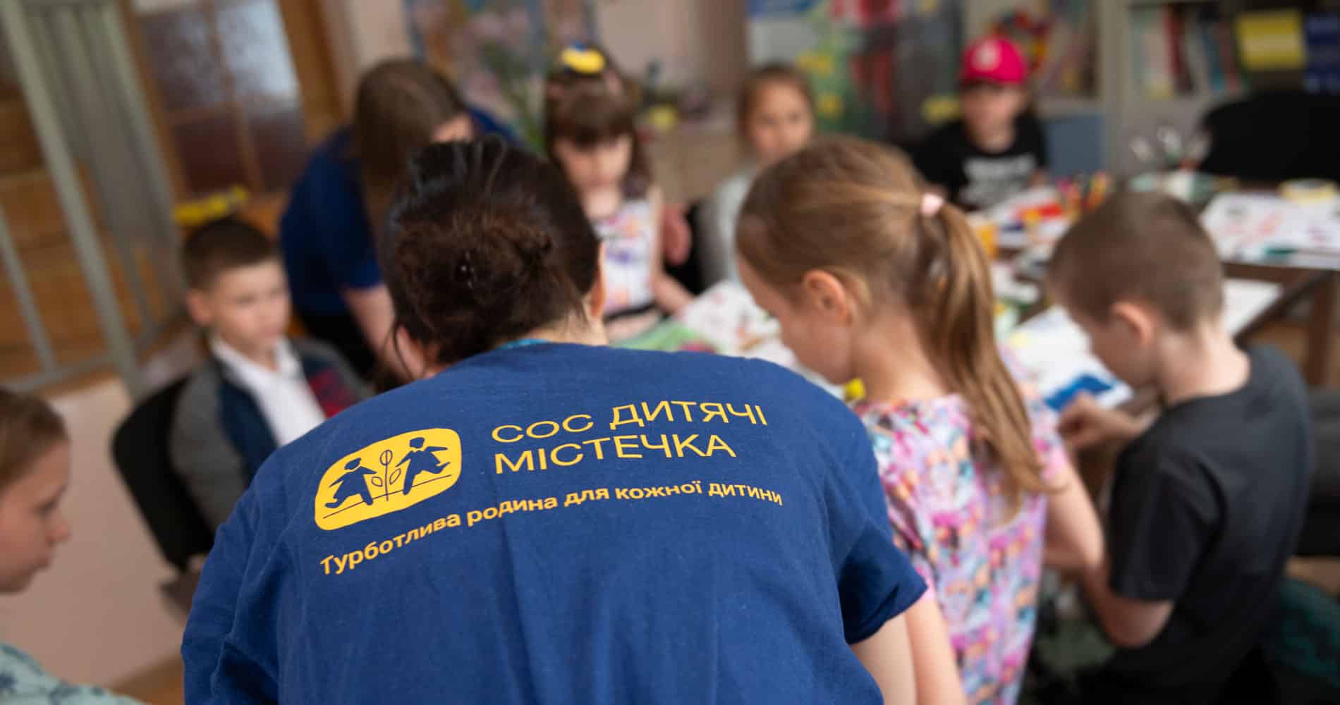 En av SOS-barnebyers sosialarbeidere, med blå t-skjorte med gul SOS-barnebyer logo, hjelper barna, de sitter rundt et bord og tegner, klipper og limer. Ukraina Foto Katerina Ilievska