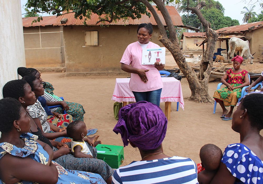 Togo: En av SOS-barnebyers medarbeidere, med rosa t-skjorte og jeans, står foran en gruppe kvinner som sitter ute blant hus lagt av leier og stein. Opplæringen de deltar på er med på å forebygge vold i hjemmet. Foto Pierre Devé