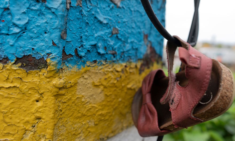 Ukraina: En murvegg malt i det ukrainske flaggets farger, på en ledning henger en liten, rosa, barnesandal. Foto Katerina Ilievska