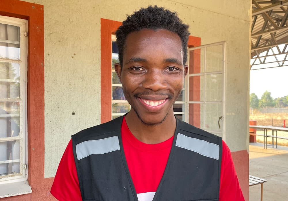 Huambo: Pedro står utenfor et hus med gule vegger og oransje vinduslister. Han smiler og har kort, mørkt hår, rød t-skjorte og sort vest med refleksfelt. Foto Turid Weisser