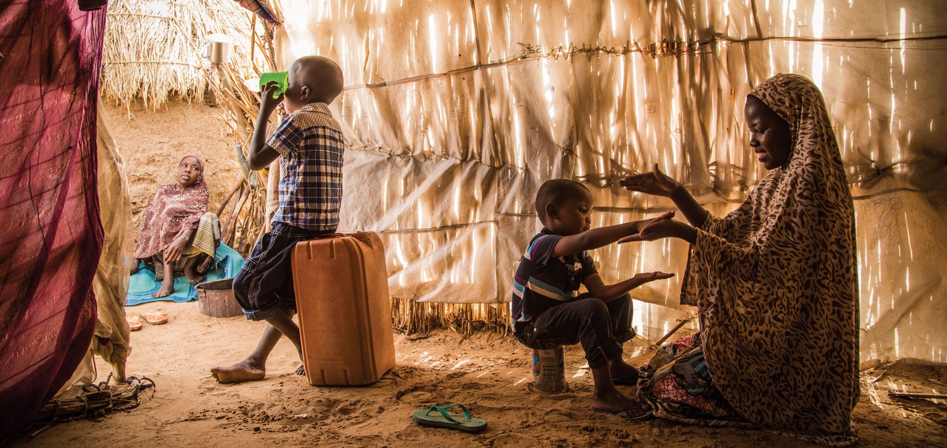Fadderskap i endring: En mor fra Niger og barna hennes sitter inne i ei hytte laget av strå. Hun leker med det minste barnet, en av sønnene sitter på ei plastkanne og drikker av en grønn kopp. Foto Vincent Tremeau