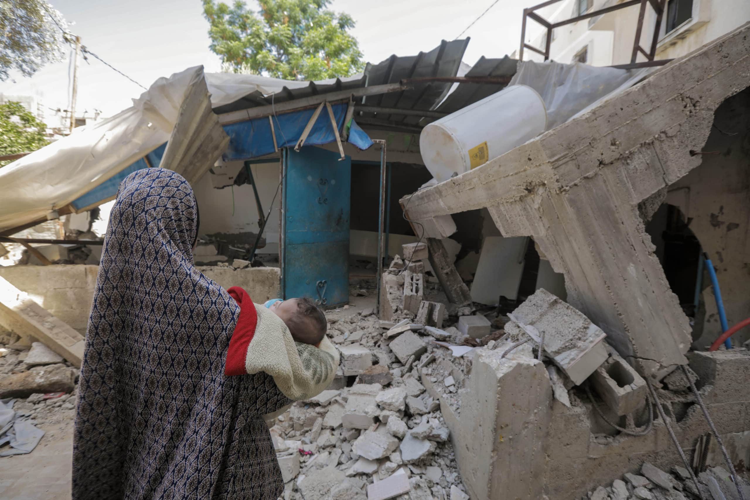 En kvinne holder en baby i armene foran et hus i ruiner i Gaza.