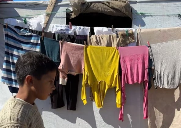 Gaza flyktningleir: Gutt med beige t-skjorte går forbi ei tørkesnor med fargerike klær, som henger mellom teltene. Foto: fra videoen/SOS-barnebyer Palestina